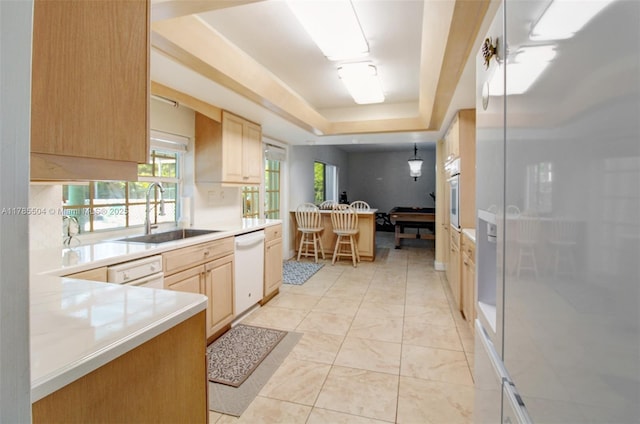 kitchen with a tray ceiling, light countertops, light brown cabinets, a sink, and white appliances