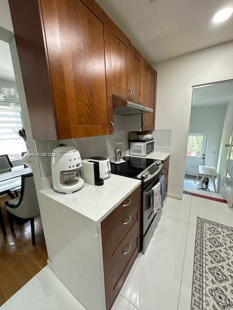 kitchen featuring under cabinet range hood, light countertops, backsplash, light tile patterned floors, and stainless steel electric stove