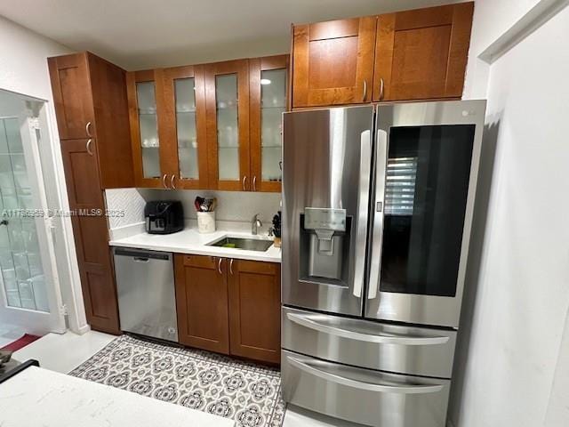 kitchen featuring a sink, glass insert cabinets, brown cabinetry, light countertops, and appliances with stainless steel finishes