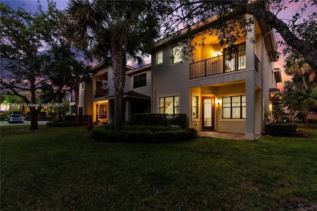 back of house at dusk featuring stucco siding, a yard, and a balcony