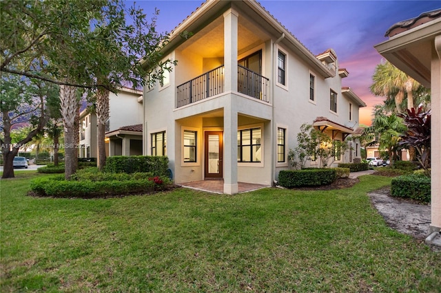back of house at dusk featuring a lawn, a balcony, and stucco siding