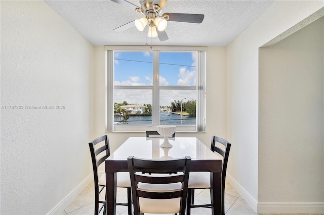 dining room featuring a water view, a ceiling fan, light tile patterned floors, baseboards, and a textured ceiling