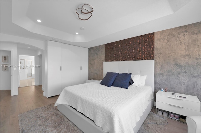 bedroom with wood finished floors, a closet, arched walkways, a raised ceiling, and recessed lighting