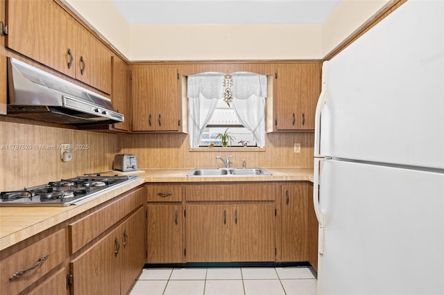 kitchen with a sink, stainless steel gas cooktop, light countertops, under cabinet range hood, and freestanding refrigerator