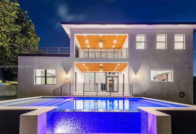 back of house at night featuring stucco siding, an outdoor pool, and a balcony