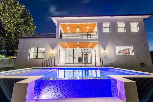 back of house at twilight with stucco siding, an outdoor pool, and a balcony