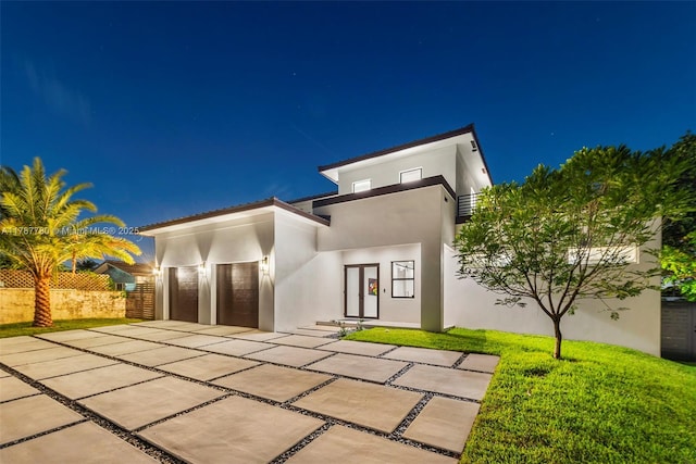 back of house at night with an attached garage, a lawn, driveway, and stucco siding