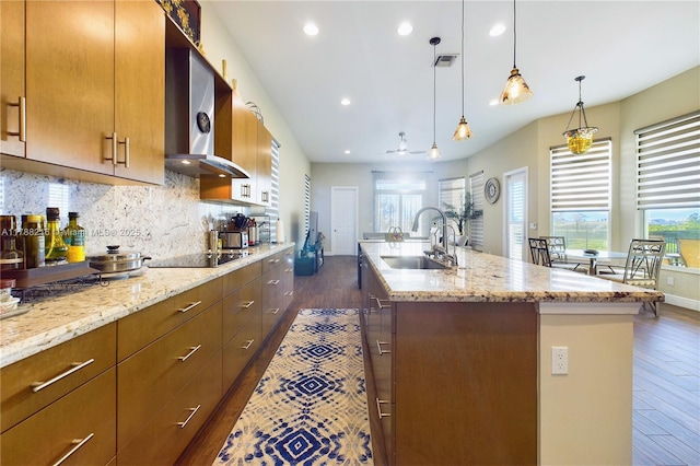 kitchen with black electric cooktop, a sink, dark wood finished floors, wall chimney range hood, and an island with sink