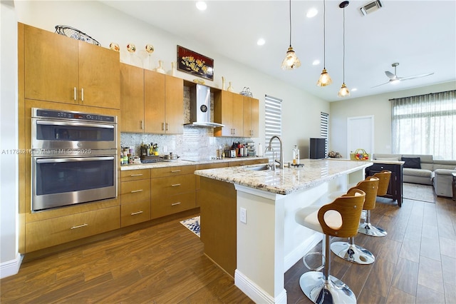 kitchen featuring stainless steel double oven, visible vents, a sink, wall chimney exhaust hood, and dark wood-style flooring
