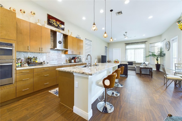 kitchen featuring dark wood-style floors, visible vents, stainless steel double oven, wall chimney range hood, and a breakfast bar