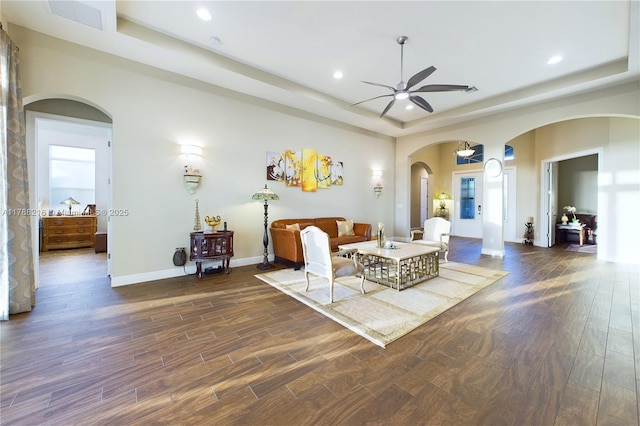 living area featuring arched walkways, dark wood finished floors, a ceiling fan, and a tray ceiling