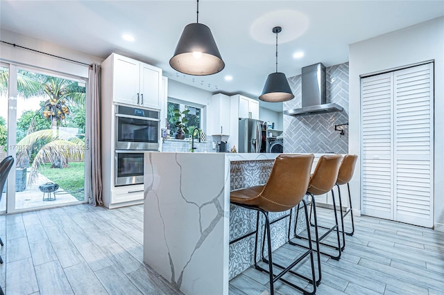 kitchen featuring tasteful backsplash, light stone counters, appliances with stainless steel finishes, white cabinetry, and wall chimney exhaust hood