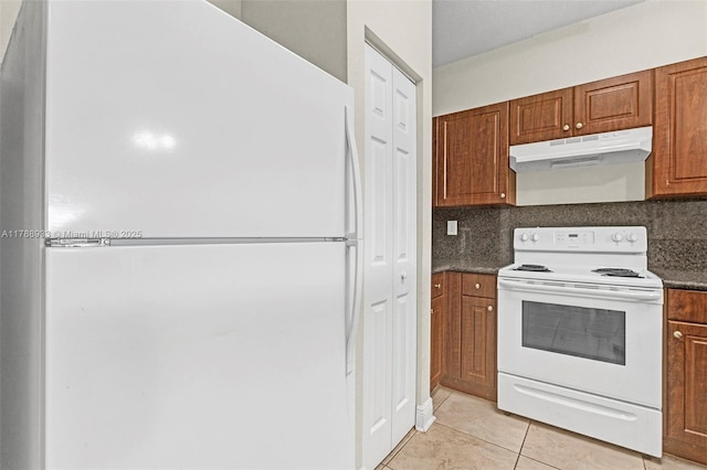 kitchen with dark countertops, brown cabinets, white appliances, light tile patterned floors, and under cabinet range hood