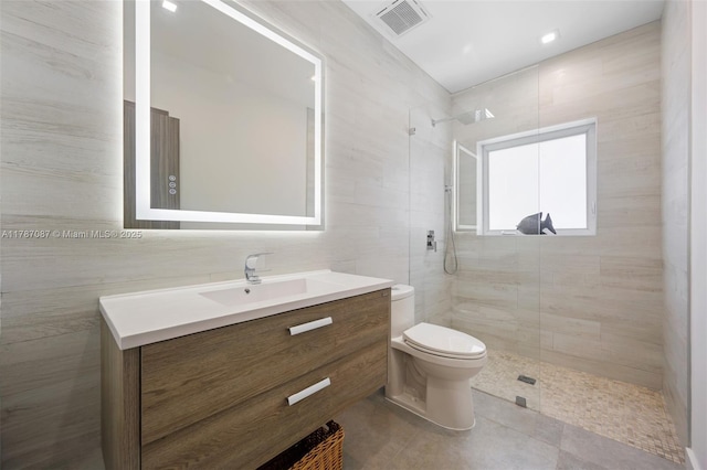 full bathroom with a tile shower, tile walls, toilet, visible vents, and vanity