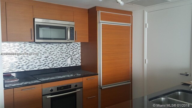 kitchen featuring brown cabinetry, appliances with stainless steel finishes, dark countertops, backsplash, and a sink