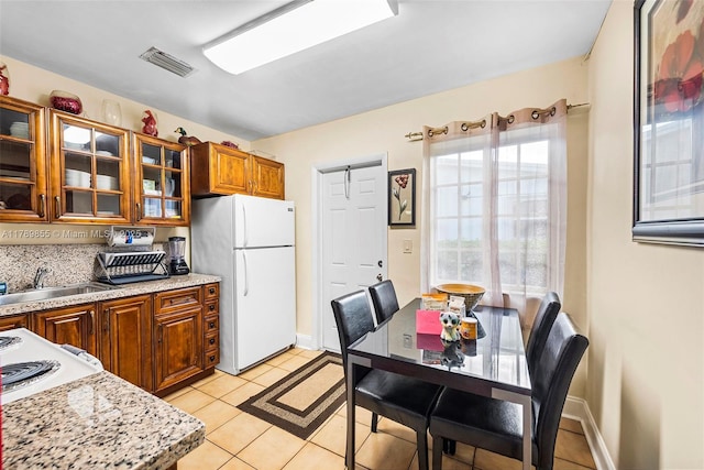 kitchen featuring brown cabinetry, freestanding refrigerator, glass insert cabinets, a sink, and light tile patterned floors