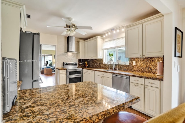 kitchen featuring a sink, stainless steel appliances, decorative backsplash, wall chimney exhaust hood, and a ceiling fan