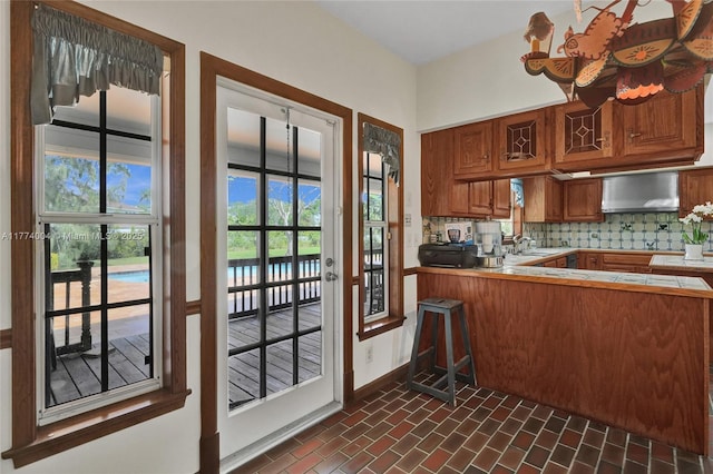 kitchen featuring wall chimney range hood, brick floor, tile counters, brown cabinetry, and baseboards
