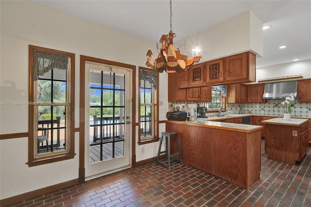 kitchen with wall chimney range hood, a notable chandelier, light countertops, brown cabinetry, and a peninsula