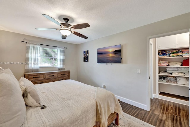 bedroom with a textured ceiling, ceiling fan, baseboards, dark wood-style flooring, and a walk in closet