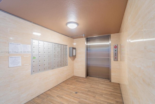 hallway with a textured ceiling, tile walls, elevator, and mail area