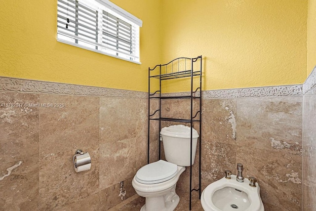 bathroom featuring toilet, a wainscoted wall, a bidet, and tile walls