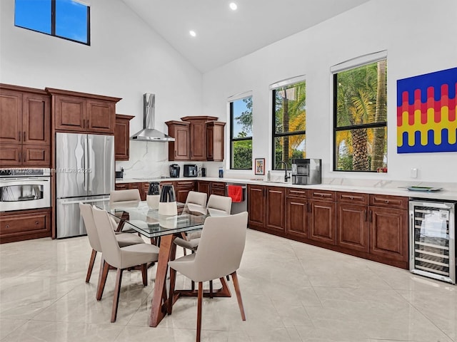 kitchen with wall chimney range hood, appliances with stainless steel finishes, light countertops, beverage cooler, and high vaulted ceiling