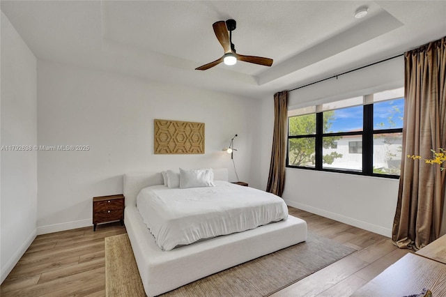 bedroom with baseboards, a ceiling fan, a tray ceiling, and light wood-style flooring