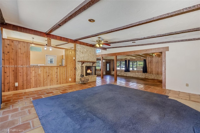 unfurnished living room with ceiling fan, a fireplace, wood walls, baseboards, and beamed ceiling
