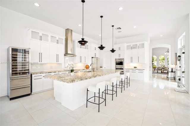 4991 E Sterling Ranch Cir, Davie, FL 33314 - kitchen with arched walkways, stainless steel double oven, wall chimney range hood, beverage cooler, and a breakfast bar