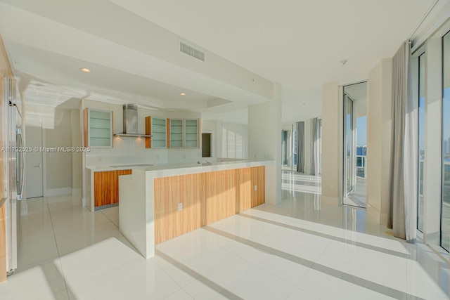 interior space with visible vents, light tile patterned flooring, glass insert cabinets, modern cabinets, and wall chimney range hood
