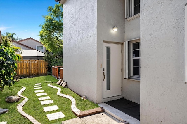 view of exterior entry with a lawn, fence, and stucco siding