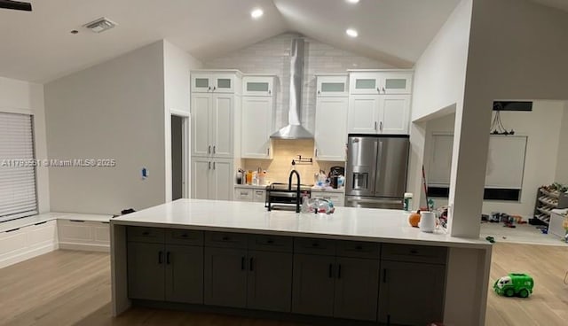 kitchen with wall chimney range hood, light wood finished floors, light countertops, white cabinetry, and stainless steel refrigerator with ice dispenser