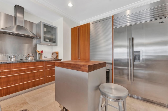 kitchen with ornamental molding, glass insert cabinets, wall chimney range hood, wood counters, and stainless steel built in refrigerator