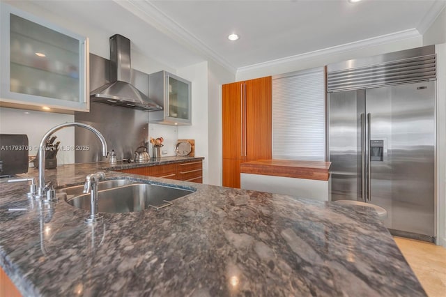 kitchen with wall chimney range hood, crown molding, a sink, built in refrigerator, and dark stone countertops