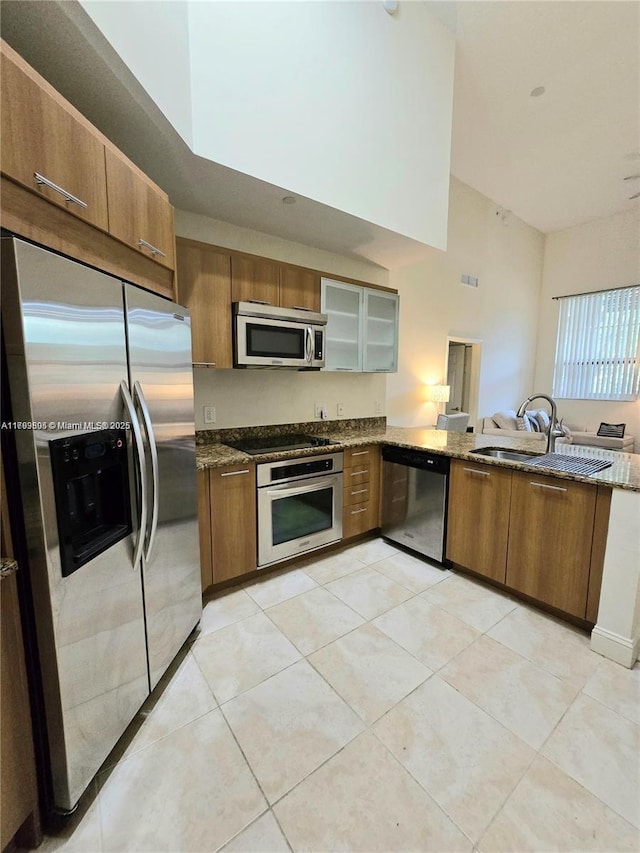 kitchen featuring brown cabinets, appliances with stainless steel finishes, a sink, stone countertops, and glass insert cabinets