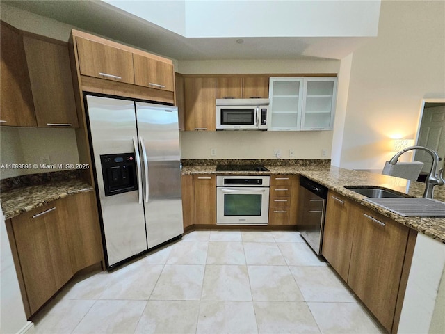 kitchen with brown cabinetry, a sink, dark stone countertops, glass insert cabinets, and stainless steel appliances