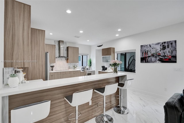 kitchen with a peninsula, modern cabinets, wall chimney range hood, a kitchen bar, and light countertops