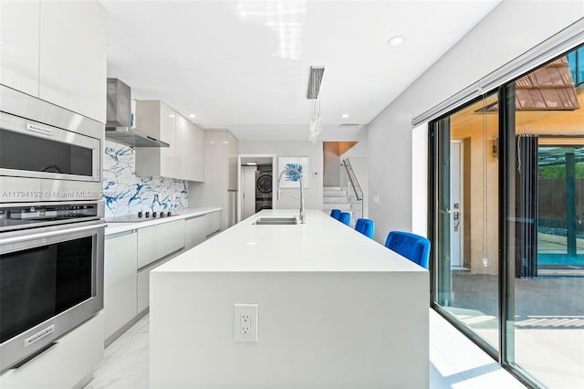 kitchen featuring wall chimney range hood, a sink, light countertops, stacked washing maching and dryer, and modern cabinets