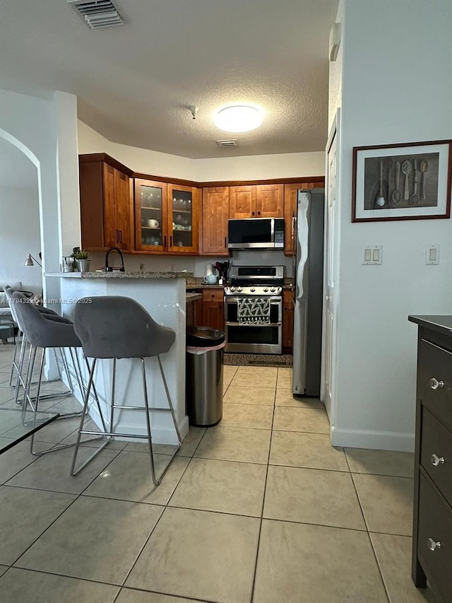 kitchen with light tile patterned flooring, brown cabinetry, stainless steel appliances, visible vents, and glass insert cabinets