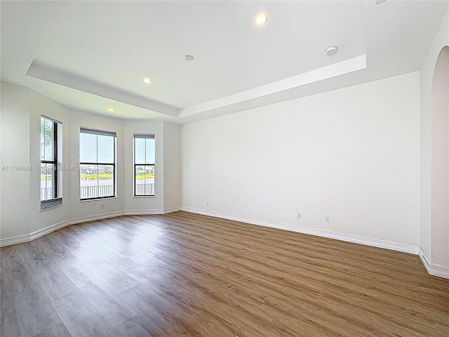empty room featuring arched walkways, wood finished floors, a raised ceiling, baseboards, and recessed lighting