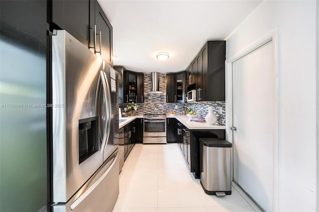 kitchen featuring light countertops, dark cabinetry, wall chimney exhaust hood, stainless steel appliances, and decorative backsplash