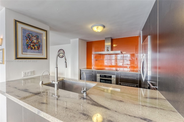 kitchen with oven, stone countertops, black electric cooktop, wall chimney exhaust hood, and a sink