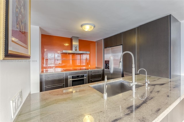 kitchen with light stone counters, a sink, wall chimney exhaust hood, stainless steel appliances, and a peninsula