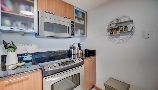 kitchen featuring brown cabinetry, glass insert cabinets, modern cabinets, dark stone countertops, and stainless steel appliances