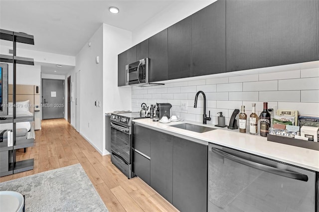 kitchen with a sink, dark cabinets, stainless steel appliances, light wood-type flooring, and backsplash