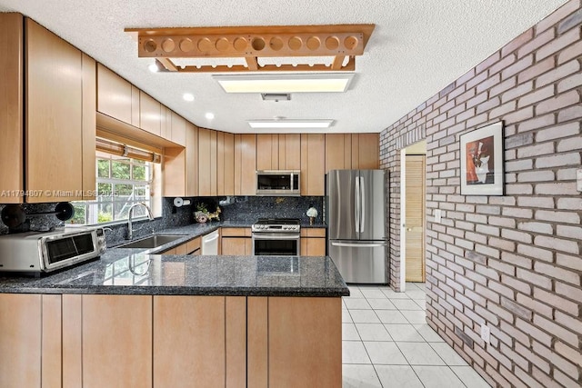 kitchen featuring a sink, a peninsula, stainless steel appliances, dark stone countertops, and brick wall