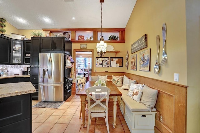 kitchen with light tile patterned flooring, a notable chandelier, stainless steel fridge, dark cabinetry, and glass insert cabinets