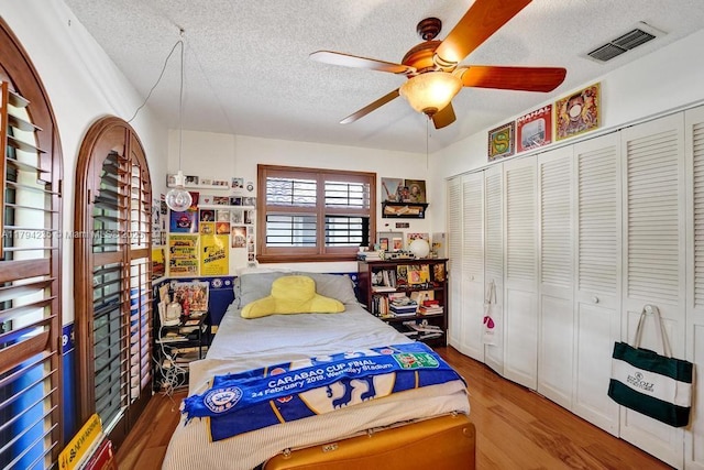 bedroom with ceiling fan, a textured ceiling, wood finished floors, visible vents, and a closet