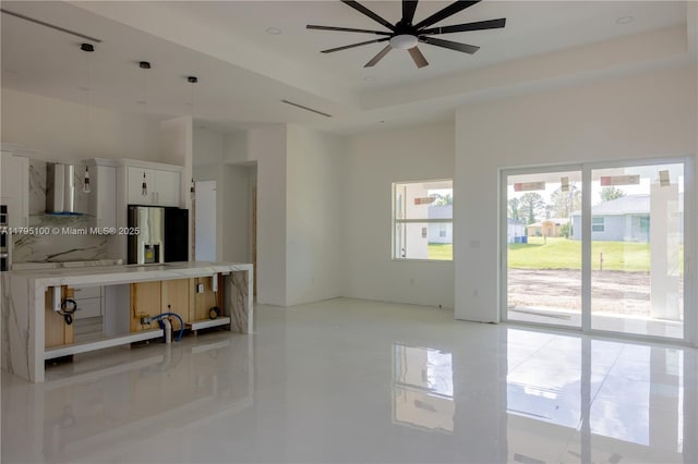 kitchen featuring wall chimney range hood, ceiling fan, stainless steel refrigerator with ice dispenser, light countertops, and white cabinetry